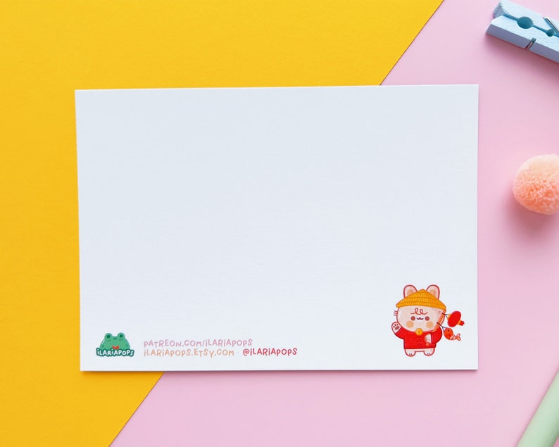 May include: A white card featuring a charming cartoon illustration of a bear dressed in a red and yellow outfit, holding a red fish. The card includes the text "PATREON.COM/ILARIAPOPS ILARIAPOPS.ETSY.COM @ILARIAPOPS" printed at the bottom.