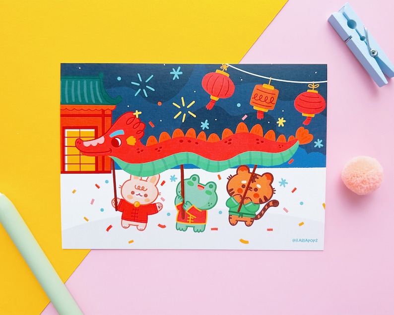 May include: A colourful illustration of three animals celebrating Chinese New Year. A red dragon puppet is held up by a rabbit, frog, and tiger. The background features a traditional Chinese building, fireworks, and red lanterns.