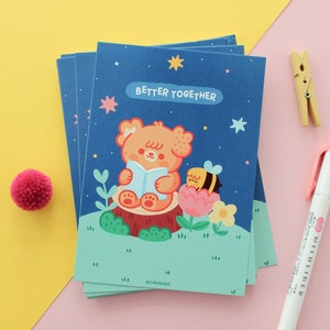 May include: A stack of cards with a blue background and a cute illustration of a bear reading a book with a bee on a flower. The text on the card says "Better Together".