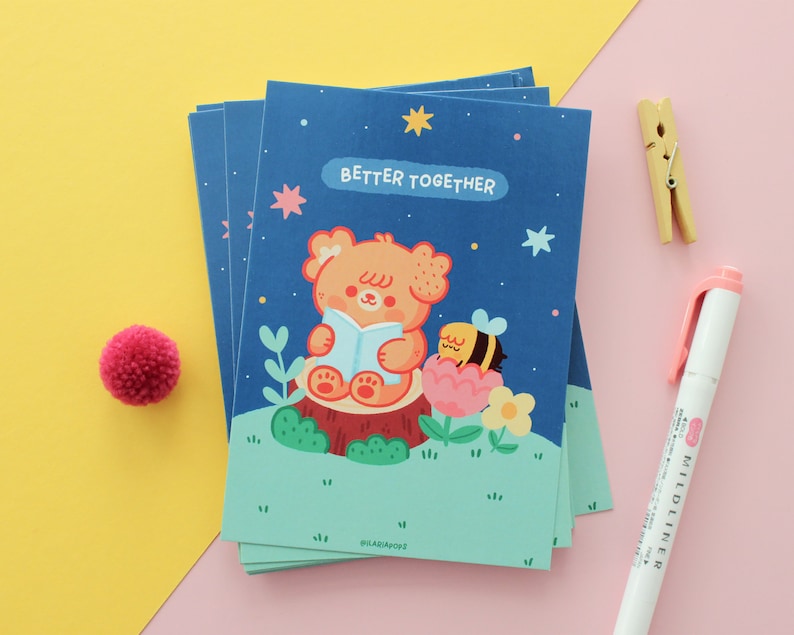 May include: A stack of cards with a blue background and a cute illustration of a bear reading a book with a bee on a flower. The text on the card says "Better Together".
