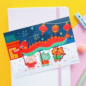 May include: A colourful illustration of a red dragon with a blue and green body, carrying four animals: a rabbit, a frog, a tiger, and a panda. The animals are wearing red and white clothing. The background is a dark blue night sky with white stars and fireworks. The illustration is on a white card with the text "#LAGADAF002" in the bottom right corner.