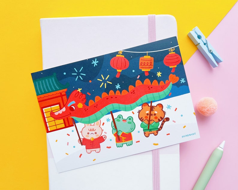 May include: A colourful illustration of a red dragon with a blue and green body, carrying four animals: a rabbit, a frog, a tiger, and a panda. The animals are wearing red and white clothing. The background is a dark blue night sky with white stars and fireworks. The illustration is on a white card with the text "#LAGADAF002" in the bottom right corner.