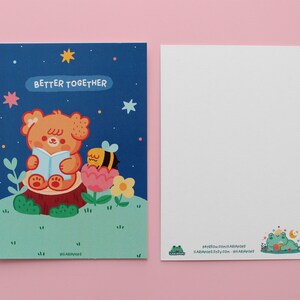 May include: A blue postcard with a cartoon illustration of a bear reading a book with a bee on a flower. The text on the postcard says "Better Together".