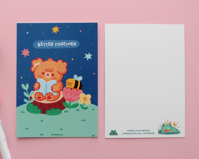 May include: A blue postcard with a cartoon illustration of a bear reading a book with a bee on a flower. The text on the postcard says "Better Together".