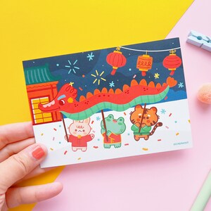 May include: A colourful illustration of a dragon dance with three animal characters: a rabbit, a frog, and a tiger. The dragon is red and green, and the characters are wearing traditional Chinese clothing. The background is a dark blue night sky with fireworks and lanterns.