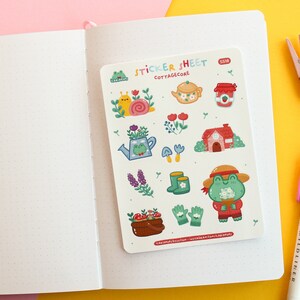 Pode incluir: Uma folha de adesivos com tema cottagecore. A folha apresenta ilustra&ccedil;&otilde;es de sapos, carac&oacute;is, flores, um bule de ch&aacute;, um pote de geleia, uma casa, botas, luvas de jardinagem e cogumelos. O texto "STICKER SHEET" e "COTTAGE CORE" est&aacute; impresso na folha.