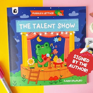 Op de afbeelding: Kinderboekcover van "The Talent Show" met een illustratie van een kikker die gitaar speelt op een podium. De titel "Puddle & Lettuce" en de tekst "Signed by the Author!" zijn ook aanwezig.