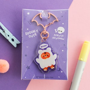 Può includere: Un portachiavi in acrilico viola e bianco con un fantasma dei cartoni animati con la scritta "Boo!" sul petto. Il portachiavi è attaccato a una catena color oro rosa e un ciondolo a forma di pipistrello. La confezione include il testo "Spooky Cute" e "Acrylic Keychain".