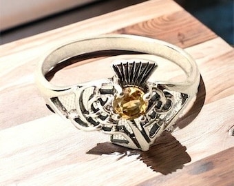 Könnte beinhalten: Ein silberfarbener Claddagh-Ring mit einem verzierten Design und einem zentralen gelben Edelstein. Der Ring liegt auf einer Holzoberfläche, die seine komplizierten Details und seine Handwerkskunst hervorhebt.