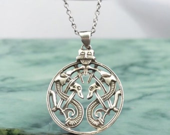 May include: A silver necklace with a circular pendant featuring an intricate design of two intertwined serpents with a stylized animal head at the top.