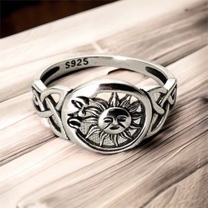 Könnte beinhalten: Ein silberner Ring mit einem Sonnen- und Monddesign. Der Ring hat eine detaillierte Sonne mit einem Gesicht in der Mitte, umgeben von Strahlen. Das Band hat ein keltisches Knotenmuster an den Seiten. Der Ring ist mit "S925" markiert.