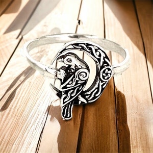 May include: A silver ring with a detailed design of a raven perched on a crescent moon. The raven and moon are surrounded by intricate Celtic knotwork.