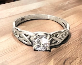 Könnte beinhalten: Ein silberner Ring mit einem keltischen Knotendesign und einem klaren, runden Stein in der Mitte.