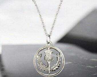Puede incluir: Un collar de plata con un colgante circular que presenta un diseño detallado y en relieve de una flor de cardo. El colgante está unido a una cadena de plata.