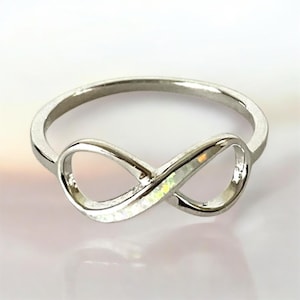 May include: A silver-colored ring featuring an infinity symbol design. The ring has a polished finish and a subtle iridescent inlay within the infinity loop. The ring is a piece of jewelry.
