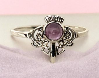 Könnte beinhalten: Ein silberner Ring mit einem Distel-Design und einem runden, lila Edelstein in der Mitte. Der Ring hat aufwendige Details um den Edelstein herum und ein schlichtes Band. Der Ring wird auf einer hellrosa Oberfläche präsentiert.