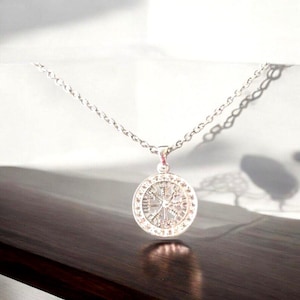May include: A silver necklace with a circular pendant featuring a detailed design of a compass with eight points. The pendant is surrounded by a circle of smaller decorative elements.