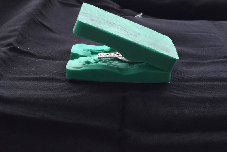 May include: A green two-piece silicone mold for casting jewelry. The mold is open and shows a silver pendant with a detailed design.