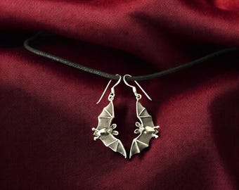 May include: A pair of silver bat earrings hanging from a black cord on a red fabric background.