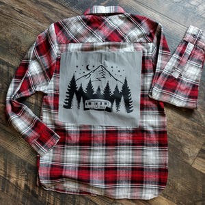 Puede incluir: Camisa de franela a cuadros rojos y blancos con un parche gris en la espalda. El parche presenta una silueta negra de una caravana, montañas y árboles bajo una luna creciente y estrellas.