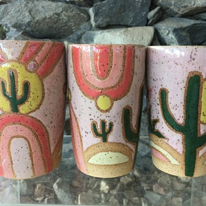 May include: Three ceramic mugs with a pink background and a desert theme. Each mug features a different cactus design with a yellow sun and pink and orange arches.