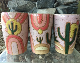 Op de afbeelding: Drie keramische mokken met een roze achtergrond en een woestijnthema. Elke mok heeft een ander cactussontwerp met een gele zon en roze en oranje bogen.