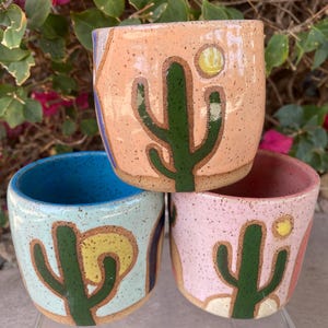 May include: Three ceramic mugs with a desert theme. Each mug features a different color background and a green cactus with a yellow sun. The mugs are stacked on top of each other.