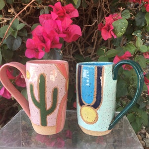 May include: Two ceramic mugs with unique designs. One mug is pink with a green cactus and a pink and orange stripe design. The other mug is light blue with a blue and black stripe design and a yellow circle.