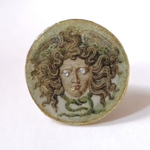 Op de afbeelding: Een ronde, ovale ring van goudkleur met een groene en bruine afbeelding van een vrouwenhoofd met slangen in haar haar. De ogen van de vrouw zijn wit en glinsteren.