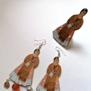 Può includere: Un paio di orecchini pendenti con un'immagine dipinta di Frida Kahlo, con perline di cornalina arancione e ciondoli d'argento. Gli orecchini sono accompagnati da un anello abbinato con un design simile.