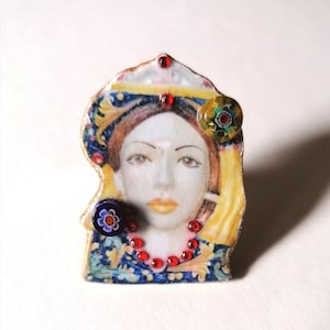 Op de afbeelding: Een kleurrijke keramische broche met het gezicht van een vrouw op een blauwe en gouden achtergrond met patronen. De broche is versierd met rode, blauwe en groene glazen kralen.