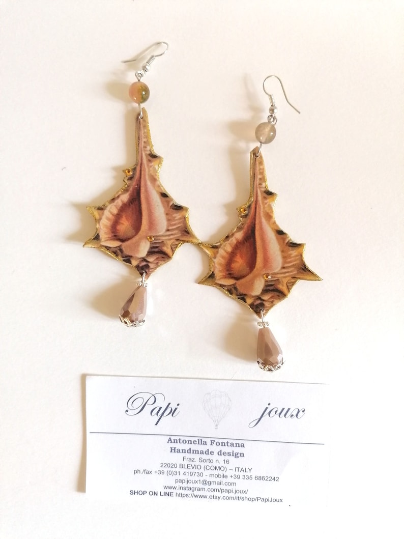 Pode incluir: Um par de brincos feitos &agrave; m&atilde;o com um design de concha, detalhes dourados e uma conta de cristal em forma de l&aacute;grima. Os brincos s&atilde;o feitos por Antonella Fontana, uma empresa de design artesanal com sede na It&aacute;lia.