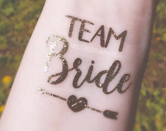 Op de afbeelding: Gouden glitter tijdelijke tattoo met de tekst "Team Bride" met daaronder een hartje en een pijl.