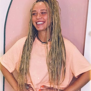 May include: A person with long, blonde dreadlocks and a peach-colored t-shirt smiles broadly. The person is wearing a black necklace and has orange nail polish. The background is a light pink color.