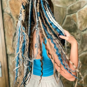 May include: A person's hair styled with a combination of dreadlocks in shades of blue, white, and brown. The dreadlocks are adorned with beads and other decorative elements. The person is wearing a blue tank top and light-colored pants.