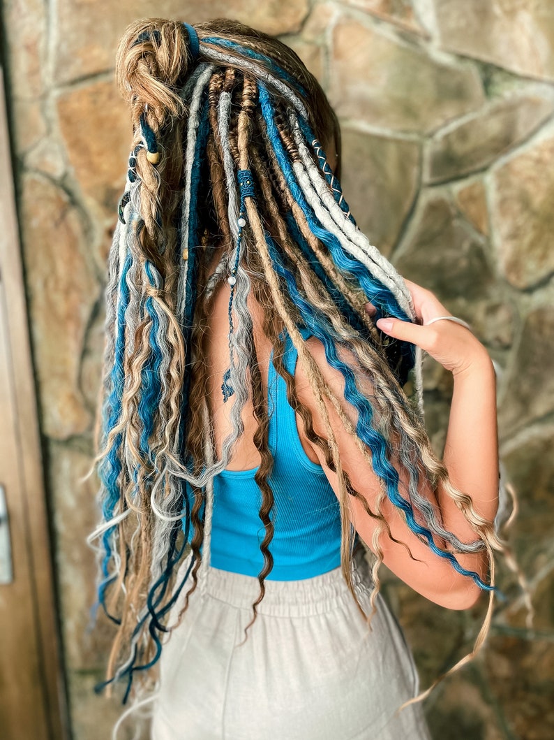 May include: A person's hair styled with a combination of dreadlocks in shades of blue, white, and brown. The dreadlocks are adorned with beads and other decorative elements. The person is wearing a blue tank top and light-colored pants.
