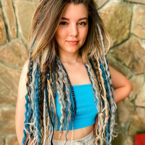 May include: A person with long, wavy hair and dreadlocks in shades of brown, blonde, gray, and blue. They are wearing a turquoise tube top and tan pants. The background is a stone wall.