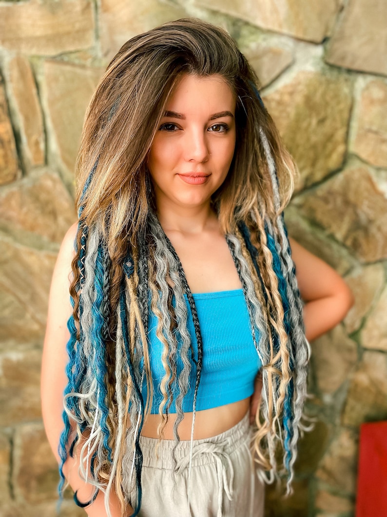 May include: A person with long, wavy hair and dreadlocks in shades of brown, blonde, gray, and blue. They are wearing a turquoise tube top and tan pants. The background is a stone wall.