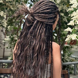 May include: Long, dark brown dreadlocks styled in a half-up, half-down look. The hair is wavy and falls down the back. The top section is tied up in a bun. The background features white flowers and greenery.