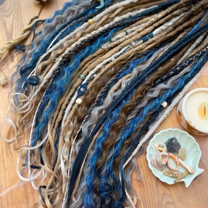 May include: A collection of handmade dreadlocks in shades of blue, brown, gray, and blonde. The dreads are adorned with beads and decorative wraps. The image is taken on a wooden surface, with a candle and seashell dish in the background.