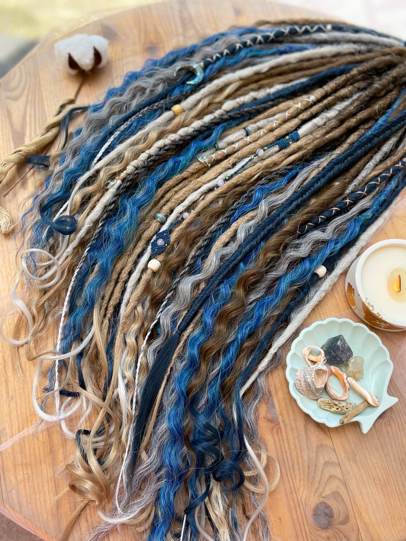 May include: A collection of handmade dreadlocks in shades of blue, brown, gray, and blonde. The dreads are adorned with beads and decorative wraps. The image is taken on a wooden surface, with a candle and seashell dish in the background.