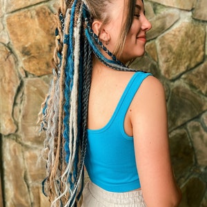 May include: A woman with long, multi-colored dreadlocks. The dreadlocks are a mix of blue, gray, and tan. She is wearing a bright blue ribbed tank top and light-colored pants. The background is a stone wall.