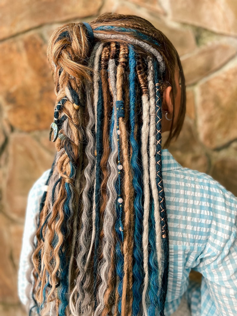 May include: A person's hair styled with a combination of dreadlocks and loose waves. The hair features a mix of colors, including brown, blue, and gray, with decorative beads and wraps. The style is worn with a blue and white checkered shirt.