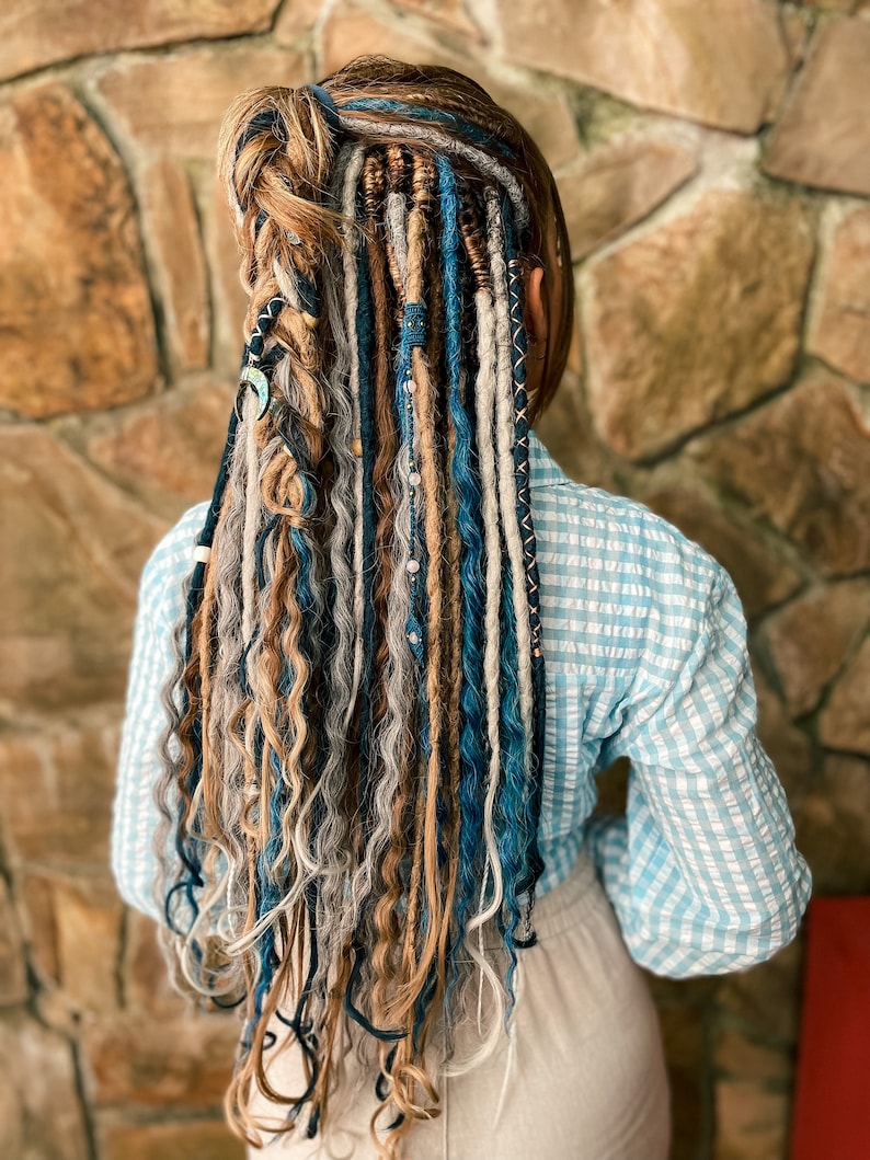 May include: A person's back is shown, with long, colorful dreadlocks. The dreadlocks are a mix of blonde, gray, and blue, with decorative beads and accents. The person is wearing a light blue and white checkered shirt.