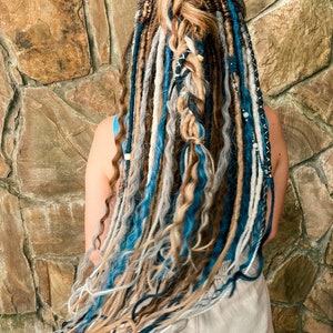 May include: A person's back view showcasing long, colorful dreadlocks. The dreadlocks are a mix of blue, gray, brown, and white, with some braided and adorned with beads. The hair is styled against a textured stone wall.