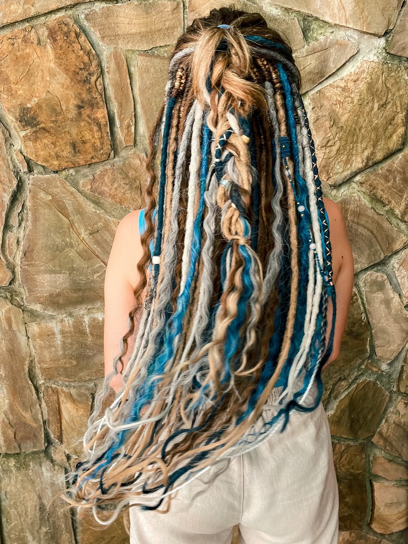 May include: A person's back view showcasing long, colorful dreadlocks. The dreadlocks are a mix of blue, gray, brown, and white, with some braided and adorned with beads. The hair is styled against a textured stone wall.