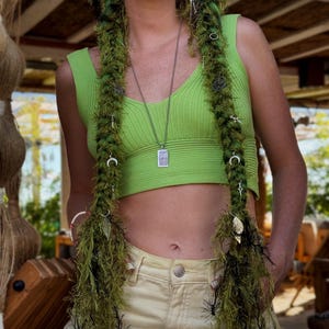 May include: A lime green ribbed crop top paired with light yellow denim jeans. Two long, textured green and black faux-fur hair extensions are draped over the shoulders, adorned with small silver charms including moons, stars, and leaves.