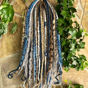 May include: A set of handmade dreadlocks in shades of blue, brown, gray, and blonde. The dreads are adorned with decorative beads and charms. The dreadlocks are hanging from a natural branch, with green foliage in the background.