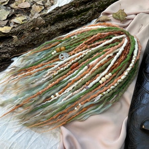 May include: A set of synthetic dreadlocks in shades of green, brown, and white with beads and charms. The dreadlocks are arranged on a light pink fabric background.