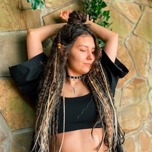 May include: A woman with long brown and blonde dreadlocks styled in a bun, wearing a black top and a black kimono-style jacket over it. She is also wearing blue jeans and has a silver necklace with a cross pendant.